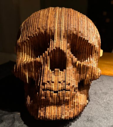 Holzskulptur eines Schädels, kunstvoll gefertigt aus gestapelten Holzstücken.